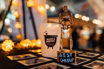 A gold skeleton trophy labeled 'Best Group' is prominently displayed on a table. Next to it, there is a white candle with the label 'Boos & Brews on the Lake.' The background features blurred, warm-toned bokeh lights adding a festive, cozy atmosphere. Several Polaroid photos are scattered on the table.