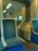 Inside a public transportation vehicle, possibly a train or subway car. The seating area is upholstered with patterned blue and teal fabric, and the interior includes metallic and plastic elements. Overhead, fluorescent lighting provides illumination. Safety signs are displayed on the walls, and there are handles for passengers to hold onto.
