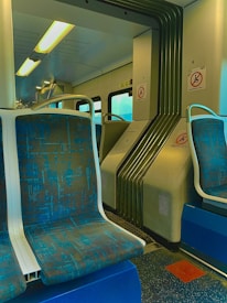 Inside a public transportation vehicle, possibly a train or subway car. The seating area is upholstered with patterned blue and teal fabric, and the interior includes metallic and plastic elements. Overhead, fluorescent lighting provides illumination. Safety signs are displayed on the walls, and there are handles for passengers to hold onto.