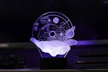 An artistic 3D representation using LED light depicts a wireframe hand holding a celestial sphere. The sphere contains abstract representations of planets, stars, and orbit paths, creating a floating cosmic scene.