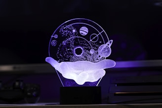 An artistic 3D representation using LED light depicts a wireframe hand holding a celestial sphere. The sphere contains abstract representations of planets, stars, and orbit paths, creating a floating cosmic scene.