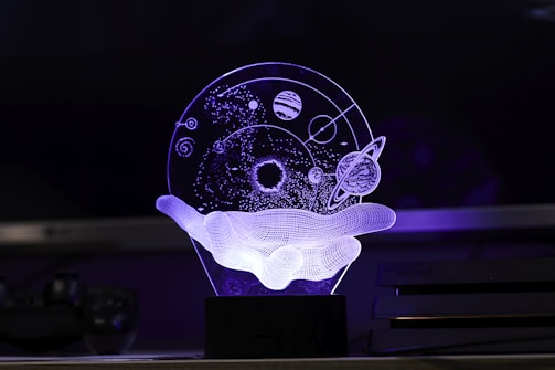 An artistic 3D representation using LED light depicts a wireframe hand holding a celestial sphere. The sphere contains abstract representations of planets, stars, and orbit paths, creating a floating cosmic scene.