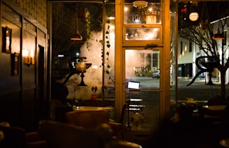 A dimly lit interior of a cozy cafe with vintage-style furnishings. Warm lighting from wall-mounted lamps illuminates the dark wooden walls. Large glass windows provide a view of the quiet street outside, with a tree and a building visible. A laptop can be seen on a small table near the entrance, and potted plants hang and climb around the windows.