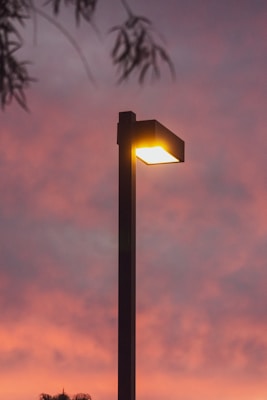 A street lamp emits a warm glow against a vibrant, colorful sky at sunset. Silhouettes of tree branches are visible in the upper corners, enhancing the ambiance.