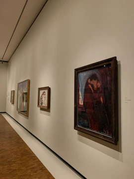 A gallery hallway with several framed paintings displayed on a beige wall. The floor is wooden, and there is soft lighting highlighting each artwork. The paintings vary in color and style, with one prominently featuring dark red and blue tones.
