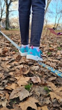 A person's legs and feet balancing on a slackline surrounded by fallen autumn leaves in a park. The shoes are colorful with blue, pink, and white accents. Trees without leaves and a bench are visible in the background under a clear sky.