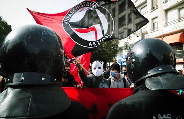 A group of people is gathered in a street protest. They are carrying a large flag with an anti-fascist emblem. Some individuals wear masks, with one wearing a distinctive white mask similar to a hockey mask, and others dressed in casual attire. Police officers in riot gear stand in the foreground, facing the group.