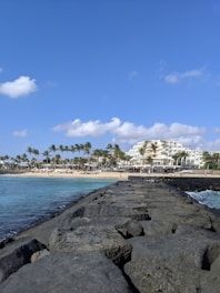 A rocky jetty extends into the ocean under a clear blue sky. Palm trees line the sandy beach on the left, with a large white resort building visible in the background. Gentle waves lap against the rocks, creating a peaceful coastal scene.