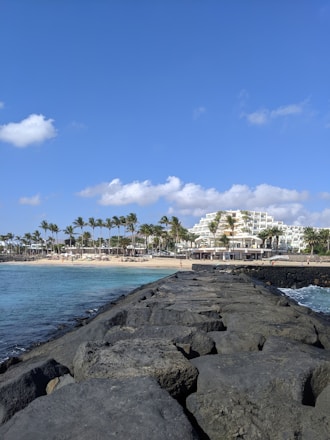 A rocky jetty extends into the ocean under a clear blue sky. Palm trees line the sandy beach on the left, with a large white resort building visible in the background. Gentle waves lap against the rocks, creating a peaceful coastal scene.