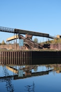 An industrial structure featuring a conveyor belt is situated near a body of water, reflecting its image clearly in the calm surface. The scene includes a signpost nearby, with greenery and a clear blue sky in the background.
