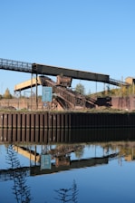 An industrial structure featuring a conveyor belt is situated near a body of water, reflecting its image clearly in the calm surface. The scene includes a signpost nearby, with greenery and a clear blue sky in the background.