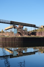 An industrial structure featuring a conveyor belt is situated near a body of water, reflecting its image clearly in the calm surface. The scene includes a signpost nearby, with greenery and a clear blue sky in the background.