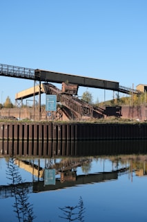 An industrial structure featuring a conveyor belt is situated near a body of water, reflecting its image clearly in the calm surface. The scene includes a signpost nearby, with greenery and a clear blue sky in the background.