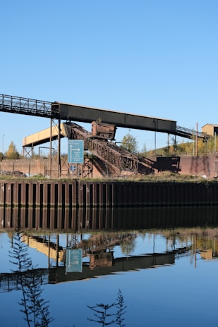 An industrial structure featuring a conveyor belt is situated near a body of water, reflecting its image clearly in the calm surface. The scene includes a signpost nearby, with greenery and a clear blue sky in the background.