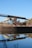 An industrial structure featuring a conveyor belt is situated near a body of water, reflecting its image clearly in the calm surface. The scene includes a signpost nearby, with greenery and a clear blue sky in the background.
