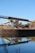An industrial structure featuring a conveyor belt is situated near a body of water, reflecting its image clearly in the calm surface. The scene includes a signpost nearby, with greenery and a clear blue sky in the background.
