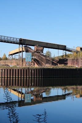 An industrial structure featuring a conveyor belt is situated near a body of water, reflecting its image clearly in the calm surface. The scene includes a signpost nearby, with greenery and a clear blue sky in the background.