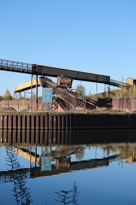 An industrial structure featuring a conveyor belt is situated near a body of water, reflecting its image clearly in the calm surface. The scene includes a signpost nearby, with greenery and a clear blue sky in the background.
