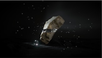 A large, rugged stone levitates in a dark environment, encircled by a metallic band. Small light particles float around, and a luminous sphere rests on the ground beneath the stone, casting a soft glow.
