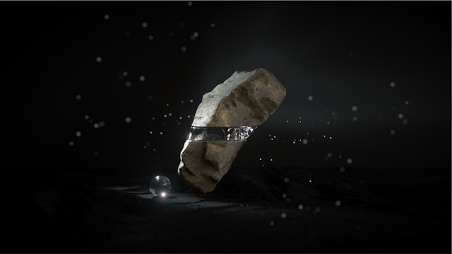 A large, rugged stone levitates in a dark environment, encircled by a metallic band. Small light particles float around, and a luminous sphere rests on the ground beneath the stone, casting a soft glow.