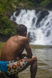 A person sits near the edge of a calm body of water, with a waterfall in the background. The individual is wearing colorful patterned shorts and appears to be contemplative, gazing towards the waterfall amidst lush greenery.