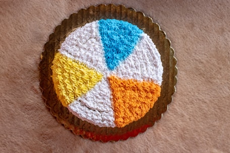 A round cake with a decorative icing design featuring segments in various colors, including blue, white, yellow, and orange, placed on a golden tray. The surface is textured with star-shaped patterns created by piped icing.