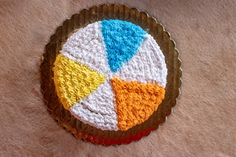 A round cake with a decorative icing design featuring segments in various colors, including blue, white, yellow, and orange, placed on a golden tray. The surface is textured with star-shaped patterns created by piped icing.