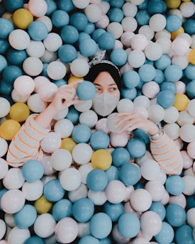 A person partially submerged in a colorful ball pit, playfully interacting with the balls. The individual is wearing a mask and a striped shirt, surrounded by an array of small, round balls in blue, pink, yellow, and white.