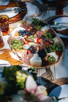 A beautifully arranged charcuterie board featuring an assortment of cheeses, meats, fruits, and nuts. Grapes, blueberries, and cherry tomatoes are artfully interspersed with slices of cured meats and various cheeses, including a wedge of blue cheese. The table setting includes elegant tableware with mustard-colored napkins, and the board is surrounded by decorative elements like woven placemats.