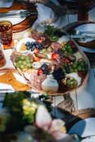 A beautifully arranged charcuterie board featuring an assortment of cheeses, meats, fruits, and nuts. Grapes, blueberries, and cherry tomatoes are artfully interspersed with slices of cured meats and various cheeses, including a wedge of blue cheese. The table setting includes elegant tableware with mustard-colored napkins, and the board is surrounded by decorative elements like woven placemats.