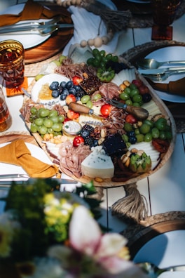 A beautifully arranged charcuterie board featuring an assortment of cheeses, meats, fruits, and nuts. Grapes, blueberries, and cherry tomatoes are artfully interspersed with slices of cured meats and various cheeses, including a wedge of blue cheese. The table setting includes elegant tableware with mustard-colored napkins, and the board is surrounded by decorative elements like woven placemats.
