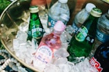 A brass container filled with several bottled beverages nestled in a bed of ice. The drinks include green glass bottles labeled Perrier and a pink Fentimans Rose Lemonade bottle, along with various plastic water bottles. The arrangement suggests a cool and refreshing setup for a picnic or outdoor event.