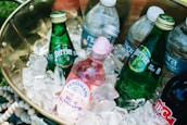 A brass container filled with several bottled beverages nestled in a bed of ice. The drinks include green glass bottles labeled Perrier and a pink Fentimans Rose Lemonade bottle, along with various plastic water bottles. The arrangement suggests a cool and refreshing setup for a picnic or outdoor event.