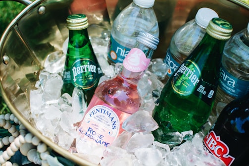 A brass container filled with several bottled beverages nestled in a bed of ice. The drinks include green glass bottles labeled Perrier and a pink Fentimans Rose Lemonade bottle, along with various plastic water bottles. The arrangement suggests a cool and refreshing setup for a picnic or outdoor event.