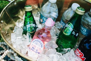 A brass container filled with several bottled beverages nestled in a bed of ice. The drinks include green glass bottles labeled Perrier and a pink Fentimans Rose Lemonade bottle, along with various plastic water bottles. The arrangement suggests a cool and refreshing setup for a picnic or outdoor event.