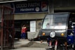 A hardware store named 'Good Faith Hardware' with a sign that includes product branding. In front of the store, there is a parked tricycle with a clear windscreen and two people inside. A person wearing a mask and colorful clothing is seen standing outside near a pile of gravel and a metal gate.
