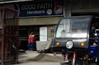 A hardware store named 'Good Faith Hardware' with a sign that includes product branding. In front of the store, there is a parked tricycle with a clear windscreen and two people inside. A person wearing a mask and colorful clothing is seen standing outside near a pile of gravel and a metal gate.