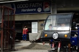 A hardware store named 'Good Faith Hardware' with a sign that includes product branding. In front of the store, there is a parked tricycle with a clear windscreen and two people inside. A person wearing a mask and colorful clothing is seen standing outside near a pile of gravel and a metal gate.