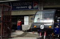 A hardware store named 'Good Faith Hardware' with a sign that includes product branding. In front of the store, there is a parked tricycle with a clear windscreen and two people inside. A person wearing a mask and colorful clothing is seen standing outside near a pile of gravel and a metal gate.