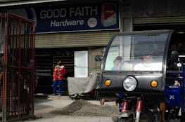 A hardware store named 'Good Faith Hardware' with a sign that includes product branding. In front of the store, there is a parked tricycle with a clear windscreen and two people inside. A person wearing a mask and colorful clothing is seen standing outside near a pile of gravel and a metal gate.
