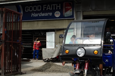 A hardware store named 'Good Faith Hardware' with a sign that includes product branding. In front of the store, there is a parked tricycle with a clear windscreen and two people inside. A person wearing a mask and colorful clothing is seen standing outside near a pile of gravel and a metal gate.
