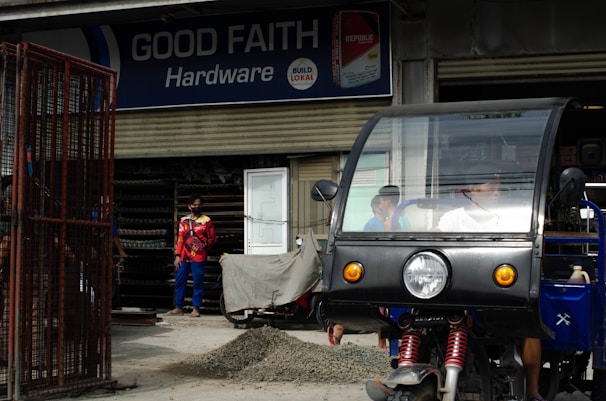A hardware store named 'Good Faith Hardware' with a sign that includes product branding. In front of the store, there is a parked tricycle with a clear windscreen and two people inside. A person wearing a mask and colorful clothing is seen standing outside near a pile of gravel and a metal gate.