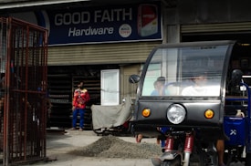 A hardware store named 'Good Faith Hardware' with a sign that includes product branding. In front of the store, there is a parked tricycle with a clear windscreen and two people inside. A person wearing a mask and colorful clothing is seen standing outside near a pile of gravel and a metal gate.