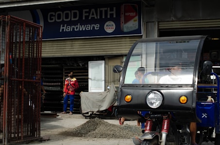 A hardware store named 'Good Faith Hardware' with a sign that includes product branding. In front of the store, there is a parked tricycle with a clear windscreen and two people inside. A person wearing a mask and colorful clothing is seen standing outside near a pile of gravel and a metal gate.