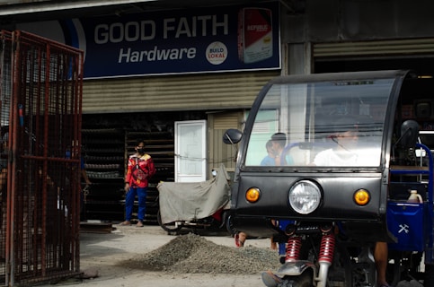 A hardware store named 'Good Faith Hardware' with a sign that includes product branding. In front of the store, there is a parked tricycle with a clear windscreen and two people inside. A person wearing a mask and colorful clothing is seen standing outside near a pile of gravel and a metal gate.