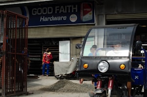 A hardware store named 'Good Faith Hardware' with a sign that includes product branding. In front of the store, there is a parked tricycle with a clear windscreen and two people inside. A person wearing a mask and colorful clothing is seen standing outside near a pile of gravel and a metal gate.