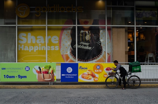 A storefront displays a large poster with 'Share Happiness' prominently featured. The poster includes a celebratory cake and festive decorations. In front of the storefront, a delivery person on a bicycle, equipped with a large green delivery bag, is preparing to deliver food. The surrounding area includes advertisements for SM Hypermarket and food items with their respective prices.