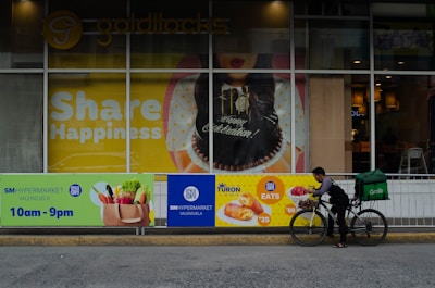A storefront displays a large poster with 'Share Happiness' prominently featured. The poster includes a celebratory cake and festive decorations. In front of the storefront, a delivery person on a bicycle, equipped with a large green delivery bag, is preparing to deliver food. The surrounding area includes advertisements for SM Hypermarket and food items with their respective prices.