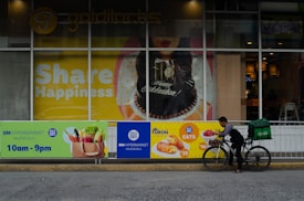 A storefront displays a large poster with 'Share Happiness' prominently featured. The poster includes a celebratory cake and festive decorations. In front of the storefront, a delivery person on a bicycle, equipped with a large green delivery bag, is preparing to deliver food. The surrounding area includes advertisements for SM Hypermarket and food items with their respective prices.