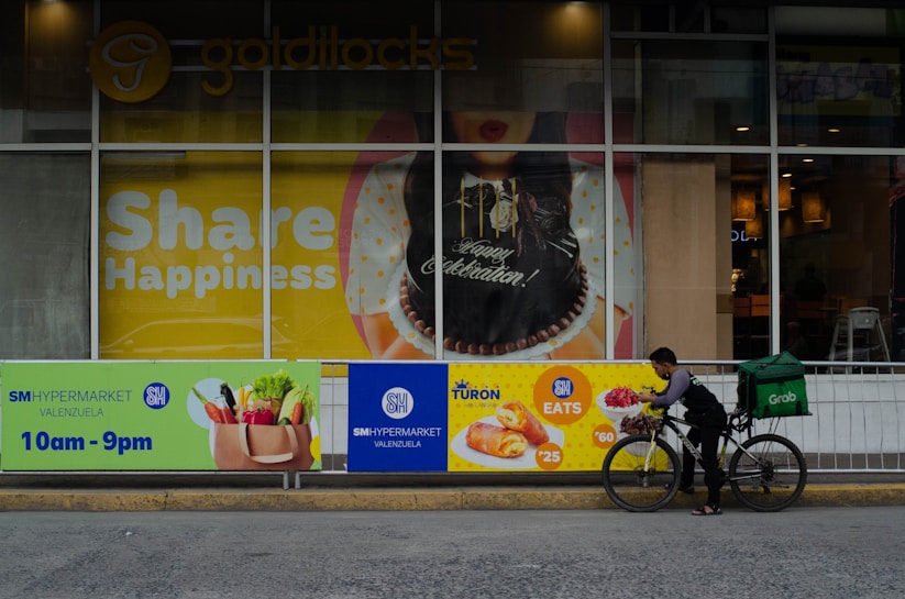 A storefront displays a large poster with 'Share Happiness' prominently featured. The poster includes a celebratory cake and festive decorations. In front of the storefront, a delivery person on a bicycle, equipped with a large green delivery bag, is preparing to deliver food. The surrounding area includes advertisements for SM Hypermarket and food items with their respective prices.